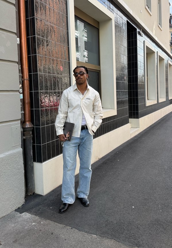 Mann med solbriller, stripete skjorte, lyse jeans og svarte sko står på fortauet og holder en bok ved siden av en bygning med svarte flislagte vegger.