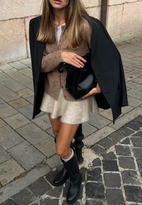 Brun strikket cardigan over en hvid top, parret med en fluffy lys beige nederdel og sorte knæhøje støvler. Stor sort håndtaske holdt i højre hånd.