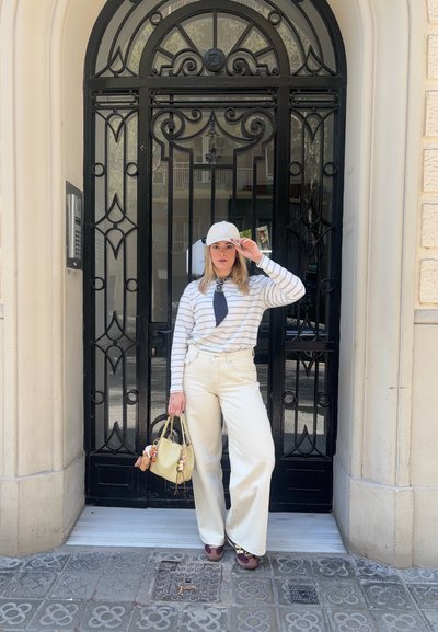 Mujer con pantalones claros, camisa de manga larga a rayas y gorra, de pie frente a una puerta ornamental de metal negro sosteniendo un bolso beige.