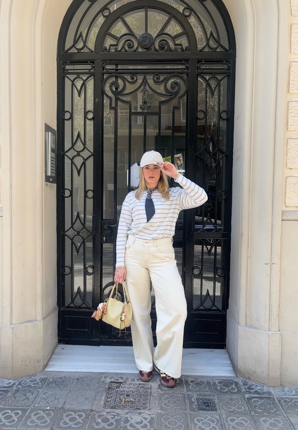 Femme en pantalon clair, chemise rayée à manches longues et casquette, debout devant une porte ornée en métal noir, tenant un sac à main beige.