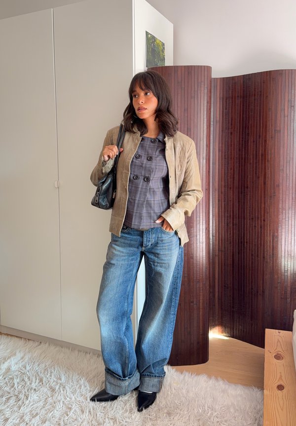 Una mujer posa en un interior con una chaqueta beige, un top de cuadros grises, unos vaqueros de pernera ancha con el bajo vuelto y zapatos negros. Lleva un bolso al hombro.