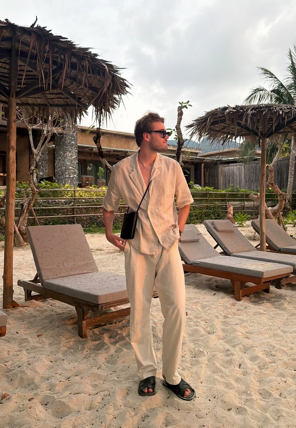 Vīrietis bēša apģērbā un saulesbrillēs stāv uz smilšainas pludmales pie atpūtas krēsliem un salmu jumta lietussargiem tropiskā kūrortā.