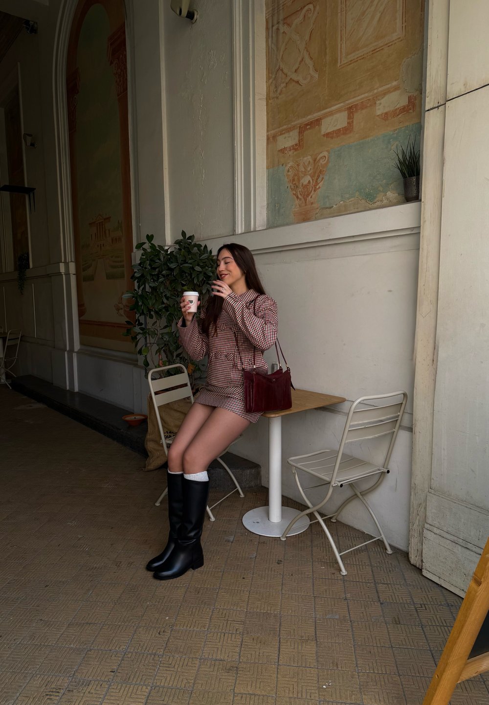 Robe à carreaux rouges et blancs, bottes noires montant jusqu'aux genoux, tenant une tasse de café, assise à côté d'une petite table et d'une plante en pot dans un cadre de café.