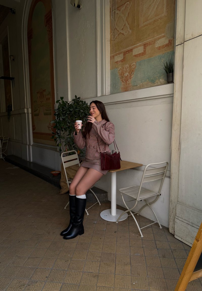 Robe à carreaux rouges et blancs, bottes noires montant jusqu'aux genoux, tenant une tasse de café, assise à côté d'une petite table et d'une plante en pot dans un cadre de café.