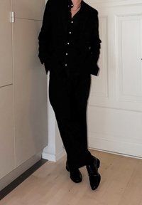 Chemise noire boutonnée avec col cranté, associée à un pantalon sombre et des chaussures noires brillantes. Texture lisse, design minimaliste.