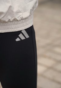 Leggings de sport noirs à texture lisse, avec un logo triangulaire gris réfléchissant sur le côté gauche et une ceinture élastique ajustée.