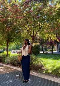 Una donna si trova accanto a alberi in fiore, indossando una maglietta bianca e una lunga gonna blu scuro, con una borsa color beige, capelli intrecciati e scarpe nere.