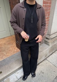 Hnědý cardigan přes černé tričko, černé široké kalhoty a lesklé černé boty. Drží průhledný kelímek se brčkem. Pozadí z kamene a dřeva.