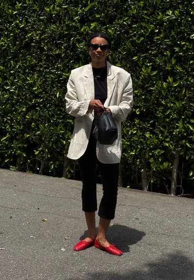 Blazer de corte amplio en beige claro sobre una blusa negra, pantalones negros recortados y bailarinas rojas de tejido. Sosteniendo un pequeño bolso negro, con un fondo de follaje verde.