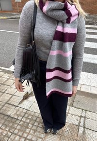 Femme debout dans la rue portant un pull gris, une large écharpe rayée gris, rose et bordeaux, un pantalon noir, un sac noir à franges et des chaussures de course.