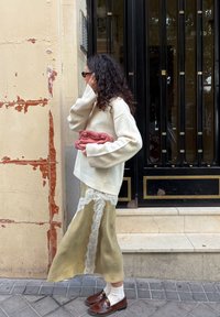 Femme aux cheveux bouclés, portant un pull crème et une jupe à bordure en dentelle, tenant un sac à main rose, debout près d'un mur usé et d'une grille en fer noir.