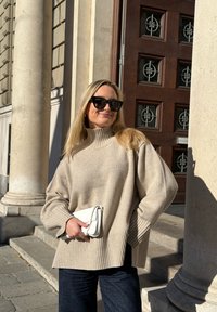 Bézs színű, túlméretezett pulóver bordázott szegéllyel, magas nyakkal és széles ujjakkal. Sötétkék farmernadrág és fehér táska. Erős kültéri megvilágítás.