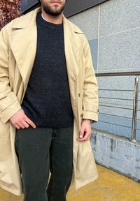 Beige trench coat sötét pulóver felett, sötétzöld nadrággal viselve. Gombokkal záródik és széles hajtókája van, textúrált anyagból készült.