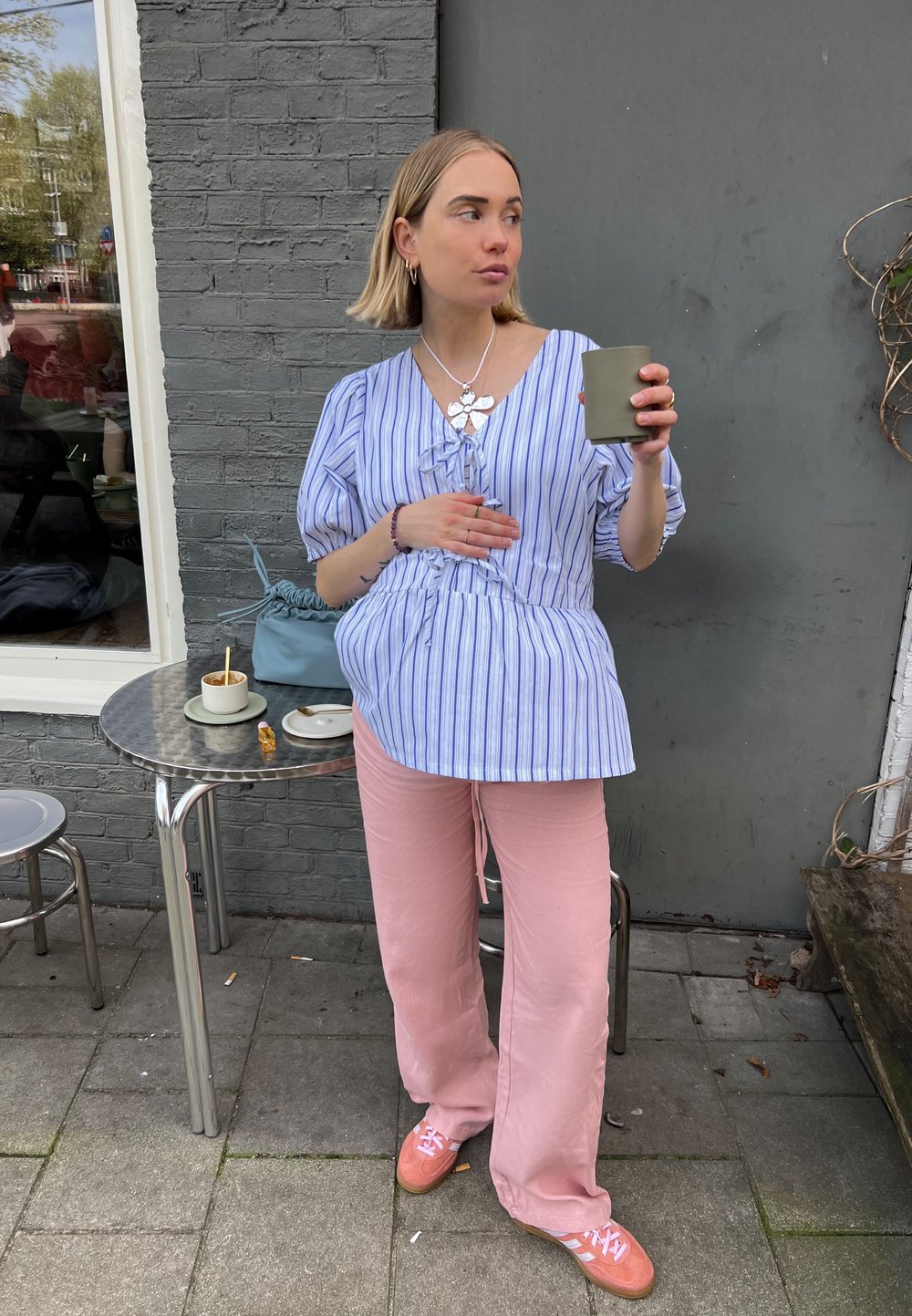 Ανοιχτό μπλε και λευκό ριγέ μπλούζα με φουσκωτά μανίκια, συνδυασμένη με χαλαρές ροζ παντελόνες. Κρατώντας ένα πράσινο κύπελλο, στέκεται μπροστά σε έναν γκρι τοίχο.
