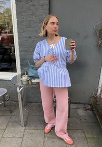 Ανοιχτό μπλε και λευκό ριγέ μπλούζα με φουσκωτά μανίκια, συνδυασμένη με χαλαρές ροζ παντελόνες. Κρατώντας ένα πράσινο κύπελλο, στέκεται μπροστά σε έναν γκρι τοίχο.