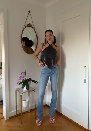 Mujer con top halter negro y jeans azules de pie junto a una mesa de vidrio y dorada con una orquídea rosa, un espejo ovalado en la pared y una puerta blanca.