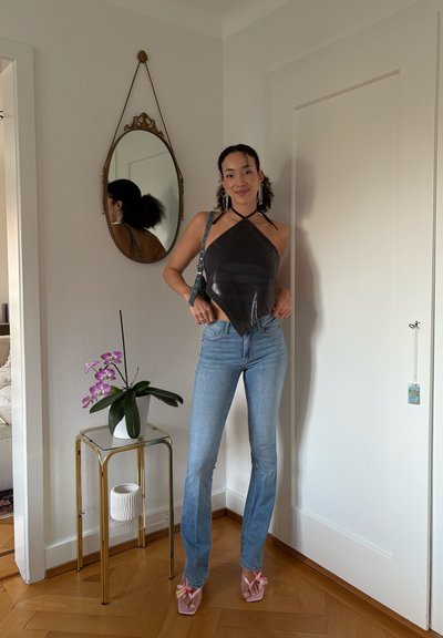 Mujer con top halter negro y jeans azules de pie junto a una mesa de vidrio y dorada con una orquídea rosa, un espejo ovalado en la pared y una puerta blanca.