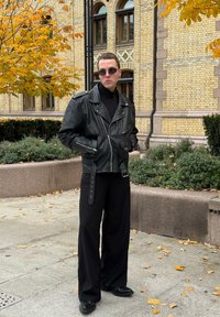 Svart läderbikerjacka med blixtlås, bärs över en svart polotröja. Lösa svarta byxor och svarta skor. Stadsmiljö med höstlöv.
