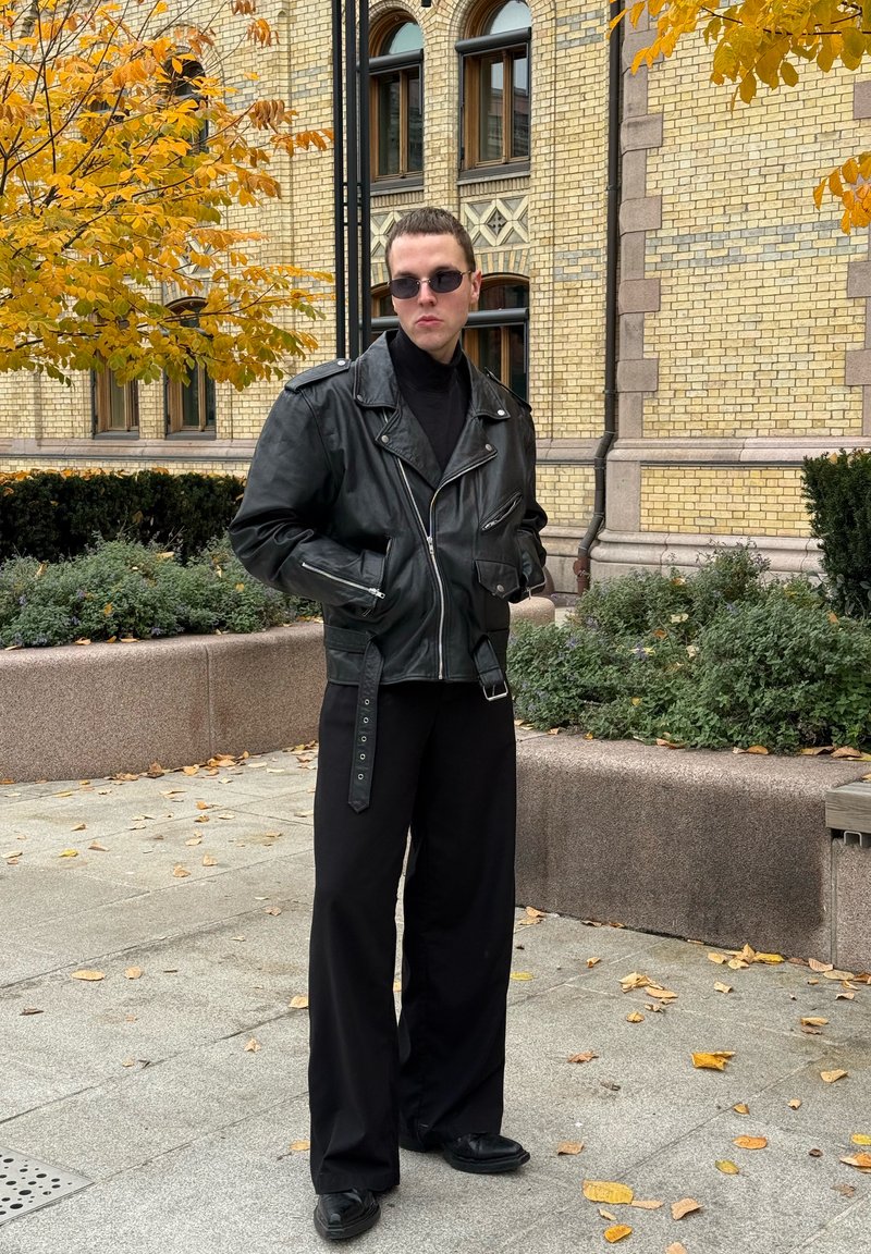 Schwarze Leder-Bikerjacke mit Reißverschlüssen, getragen über einem schwarzen Rollkragenpullover. Lockere schwarze Hose und schwarze Schuhe. Urbanes Umfeld mit herbstlichen Blättern.