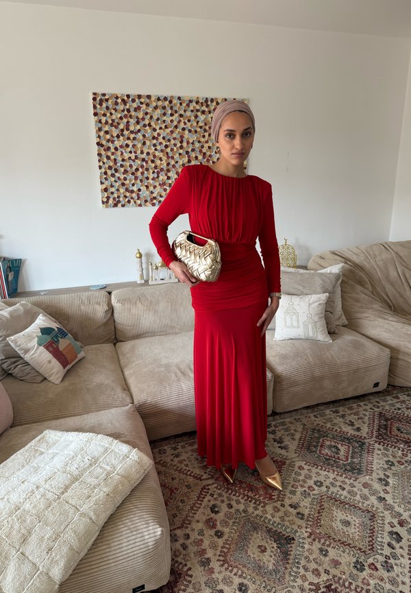 Femme en longue robe rouge et foulard beige debout dans un salon, tenant une pochette tissée dorée, avec des canapés beiges et un tapis à motifs.