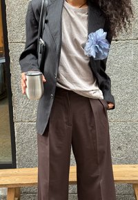 Stribet sort blazer, løse brune bukser, grå skjorte, blå stofblomst som tilbehør og en rejsekrus i rustfrit stål i hånden.