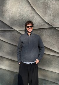 Jeune homme portant des lunettes de soleil et une veste zippée gris foncé, debout devant un mur abstrait gris texturé avec des motifs pliés.