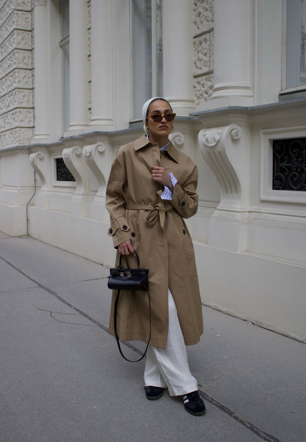 Beige trench coat med bælte, med en klassisk krave, iført over en blå og hvid stribet skjorte og hvide bukser. Sort tote taske, sorte sko.