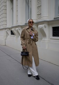 Beige trench coat med bælte, med en klassisk krave, iført over en blå og hvid stribet skjorte og hvide bukser. Sort tote taske, sorte sko.