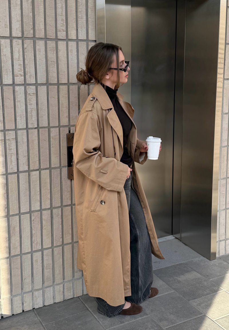 Donna con trench beige e occhiali da sole che tiene una tazza di caffè in mano, in piedi vicino a porte chiuse dell'ascensore su pavimento piastrellato.