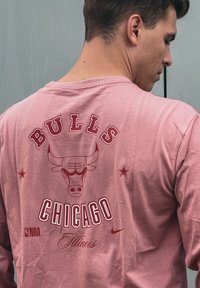 Ροζ Τ-shirt με μακριά μανίκια που διαθέτει μεγάλο κόκκινο κείμενο "BULLS" και λογότυπο ταύρου στην πλάτη, με τις λέξεις "CHICAGO" και "Illinois" από κάτω. Παρόν το λογότυπο της Nike.