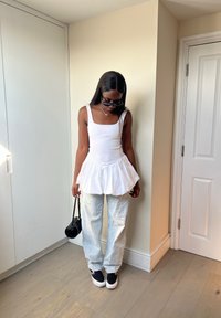 Biely peplum top s tenkými ramienkami, skombinovaný s svetlomodrými voľnými džínsami a čiernymi teniskami. Malá čierna kabelka visí z jedného ramena.