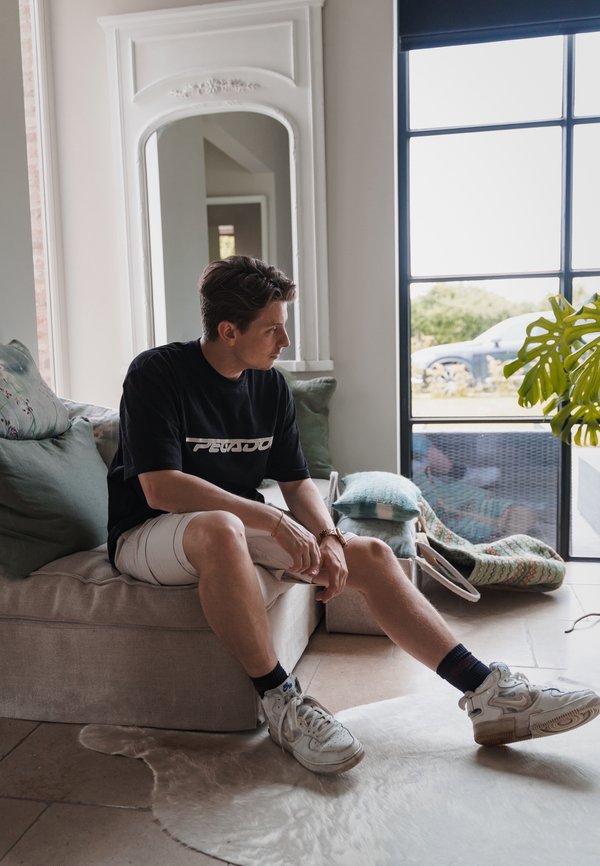 Sofá de tela gris con cojines verdes, una persona con una camiseta negra y pantalones cortos blancos sentada. Suelo claro, ventana con vista a la vegetación exterior.