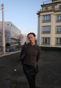 Jeune femme aux cheveux bouclés, veste rayée et jupe en cuir, debout à l'extérieur près de bâtiments résidentiels au crépuscule.