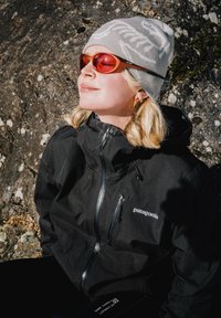 Veste imperméable noire avec une fermeture éclair, bonnet en tricot gris avec un logo, et lunettes teintées orange. L'arrière-plan présente une surface rocheuse.