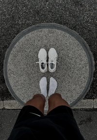 Białe buty sportowe na szarej, okrągłej platformie, widoczne białe skarpetki. W tle ciemny żwir i detale betonowe.