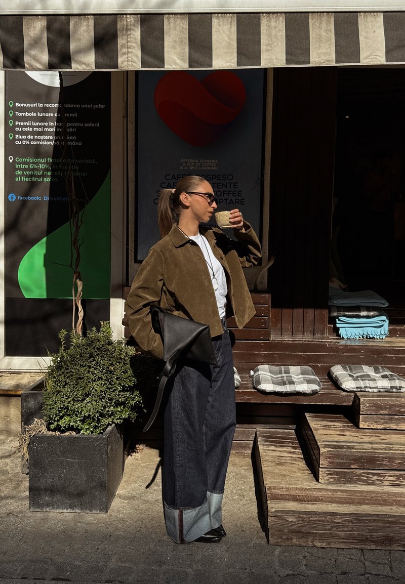 Femme en veste marron et jean large boit dans une tasse à l'extérieur d'un café avec des marches en bois et des sièges rembourrés.