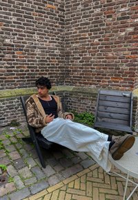 Brązowa kurtka z sztucznego futra z kapturem, czarny top bez rękawów, białe szerokie spodnie, siedząca na czarnym drewnianym krześle przy ceglanej ścianie. Widoczne zieleń.