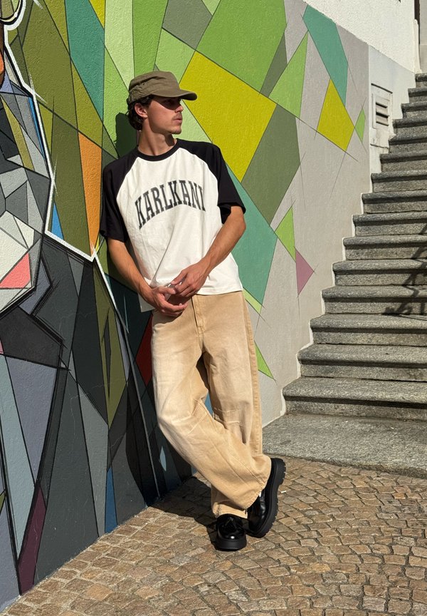 Young man wearing a cap, white and black shirt, and beige pants leaning against a colorful geometric mural next to stone stairs outdoors.