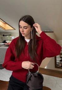 Mujer con cabello largo y castaño, vestida con un suéter rojo de punto y pendientes largos y rectangulares, sosteniendo un pequeño bolso negro en el interior.