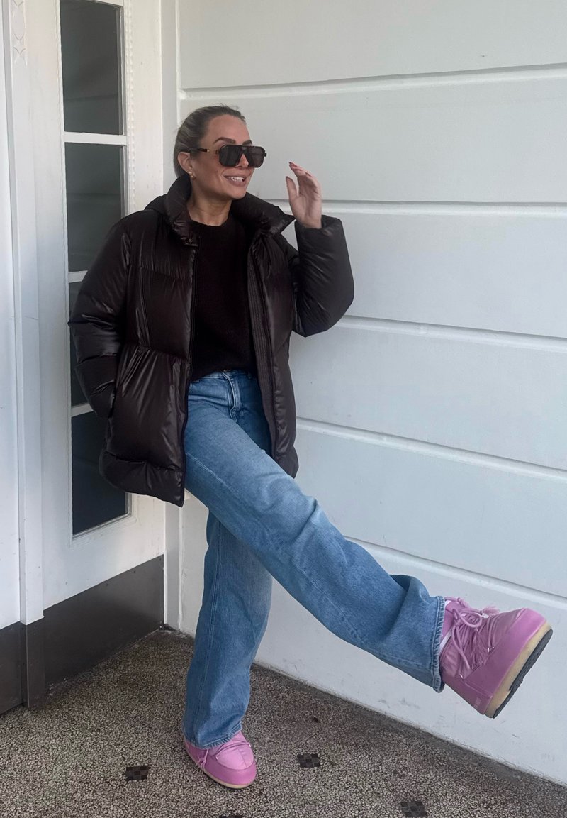 Doudoune marron, pull foncé, jean bleu clair et baskets à plateforme roses. Debout contre un mur blanc avec un cadre de porte.