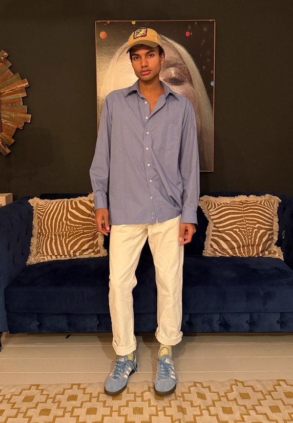 Giovane uomo con cappellino beige, camicia azzurra con bottoni, pantaloni bianchi e scarpe da ginnastica blu in piedi davanti a un divano blu scuro con cuscini decorati.