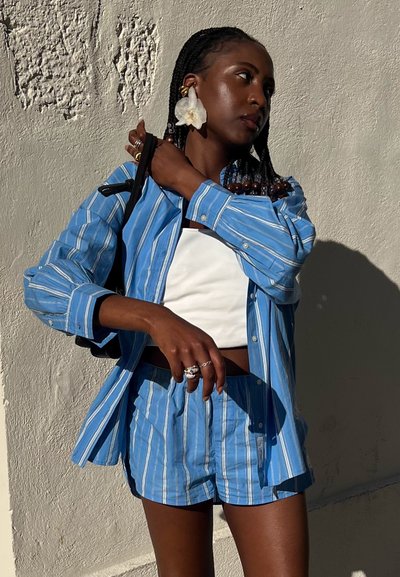 Camisa de botones a rayas azul y blanca combinada con pantalones cortos a juego. Presenta una camiseta corta blanca debajo y grandes pendientes florales.