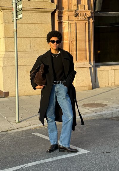 Abrigo largo negro sobre un suéter negro, jeans rectos de mezclilla azul claro y botas de tobillo. Sosteniendo un bolso marrón. Las gafas de sol acentúan el look.