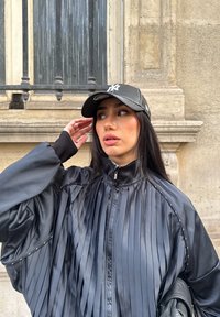 Ung kvinna som bär en svart plisserad jacka med dragkedja och en svart basebollkeps, rör vid sin keps, framför en stenbyggnad med ett fönster.