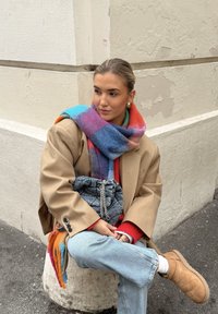 Beige oversized blazer, fargerik rutet skjerf, rød genser, blå jeans, brune støvler, grå veske med kjede, sittende på en betongblokk.
