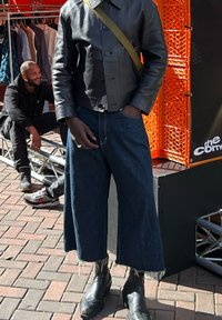 Sort læder cropped jakke med knapper, blå vidde denim jeans med flokerede kanter og sorte ankelstøvler på en murstensbaggrund.