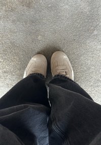 Grijze suède sneakers met platte veters en een wit logo aan de zijkant, gecombineerd met zwarte wijde broek, bekeken van bovenaf.