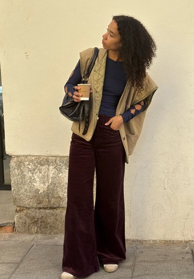 Mujer con cabello rizado, vestida con una blusa azul marino, un chaleco sin mangas beige y pantalones de pierna ancha burdeos, sosteniendo una taza de café y un bolso negro, de pie al aire libre.