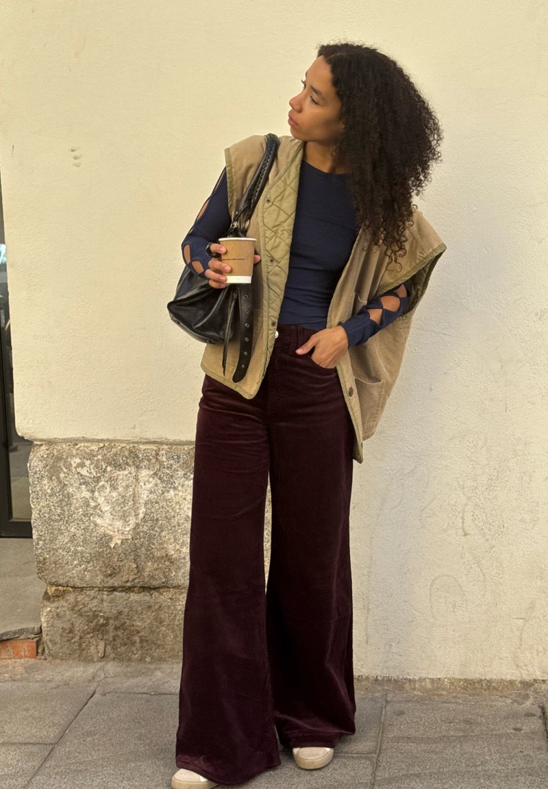 Mujer con cabello rizado, vestida con una blusa azul marino, un chaleco sin mangas beige y pantalones de pierna ancha burdeos, sosteniendo una taza de café y un bolso negro, de pie al aire libre.