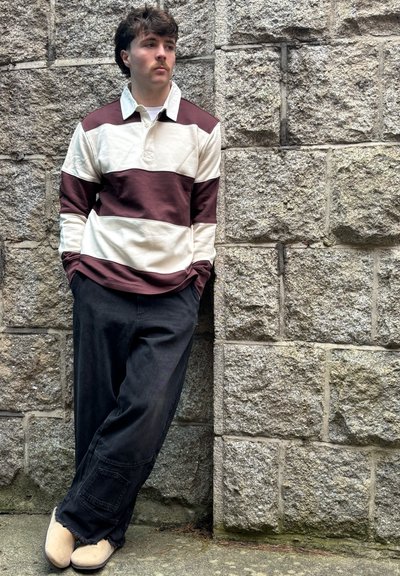 Camisa de manga larga a rayas en crema y burdeos, con cuello y tapeta frontal. Combinada con pantalones negros holgados y zapatos beige.